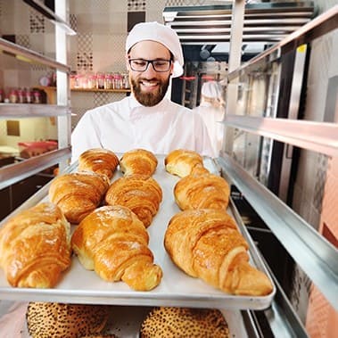 handsome male baker holds a tray with French croissants in front of a bakery and smiles.